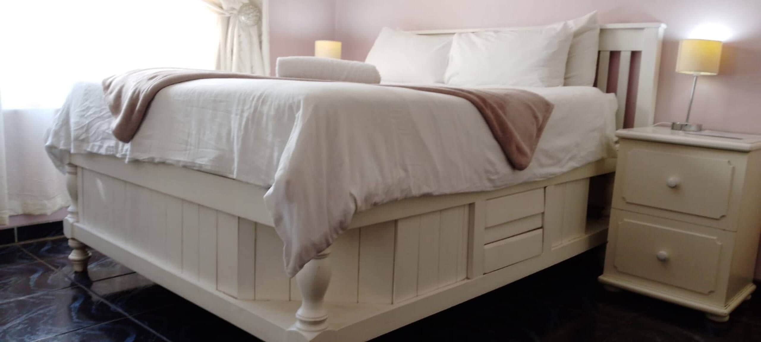 Una cama con ropa blanca y un plaid marrón, una mesita de noche con una lámpara y un mueble con cajones.