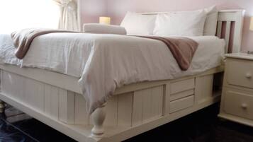 Una cama con ropa blanca y un plaid marrón, una mesita de noche con una lámpara y un mueble con cajones.