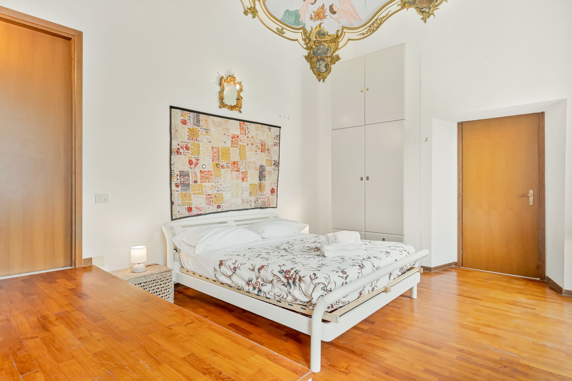 Een slaapkamer met een houten vloer, een bed met bloemenbeddels, een aan de muur hangend sprei, een kledingkast en een deur.