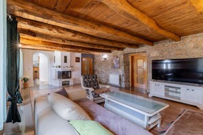 A cozy living room with a stone wall, wooden ceiling beams, a fireplace, a sofa, a television, and a kitchen area in the background. - Hilly Stone Villa for 10 Guests in Donji Vinjani (Donji Vinjani)