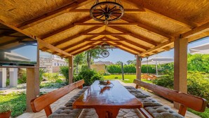 An outdoor wooden pergola with a table and benches, surrounded by greenery and a stone pathway.