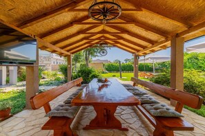 An outdoor wooden pergola with a table and benches, surrounded by greenery and a stone pathway.