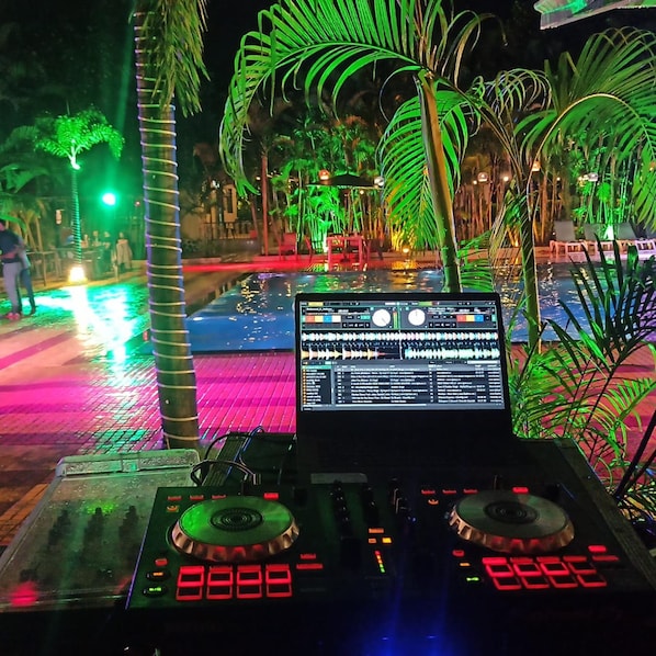 A DJ setup with a laptop and two turntables, surrounded by palm trees and a pool.
