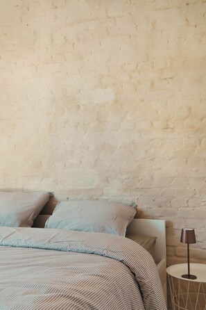 Cama com roupa de cama listrada em frente a uma parede texturizada, luminária de cabeceira e uma pequena mesa redonda.