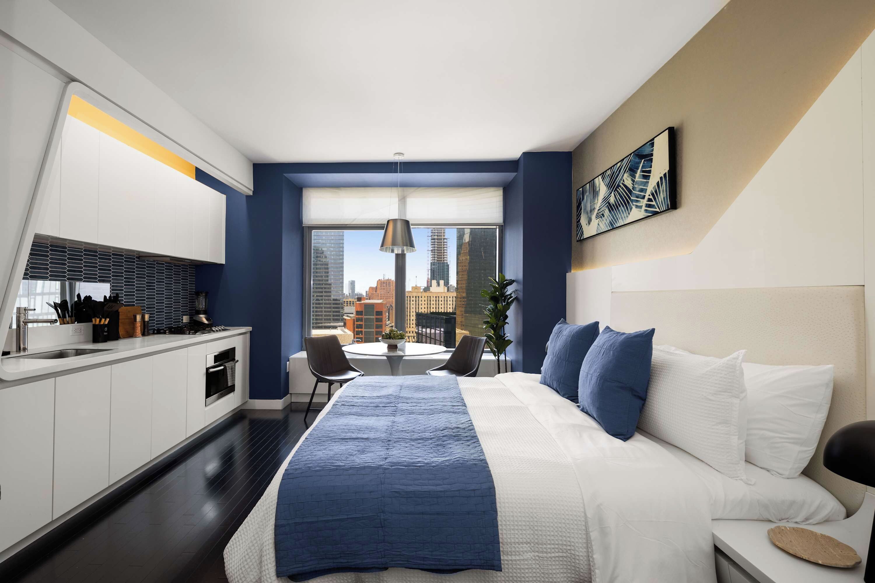 A modern hotel room with a large bed, a dining area, and a view of the cityscape.