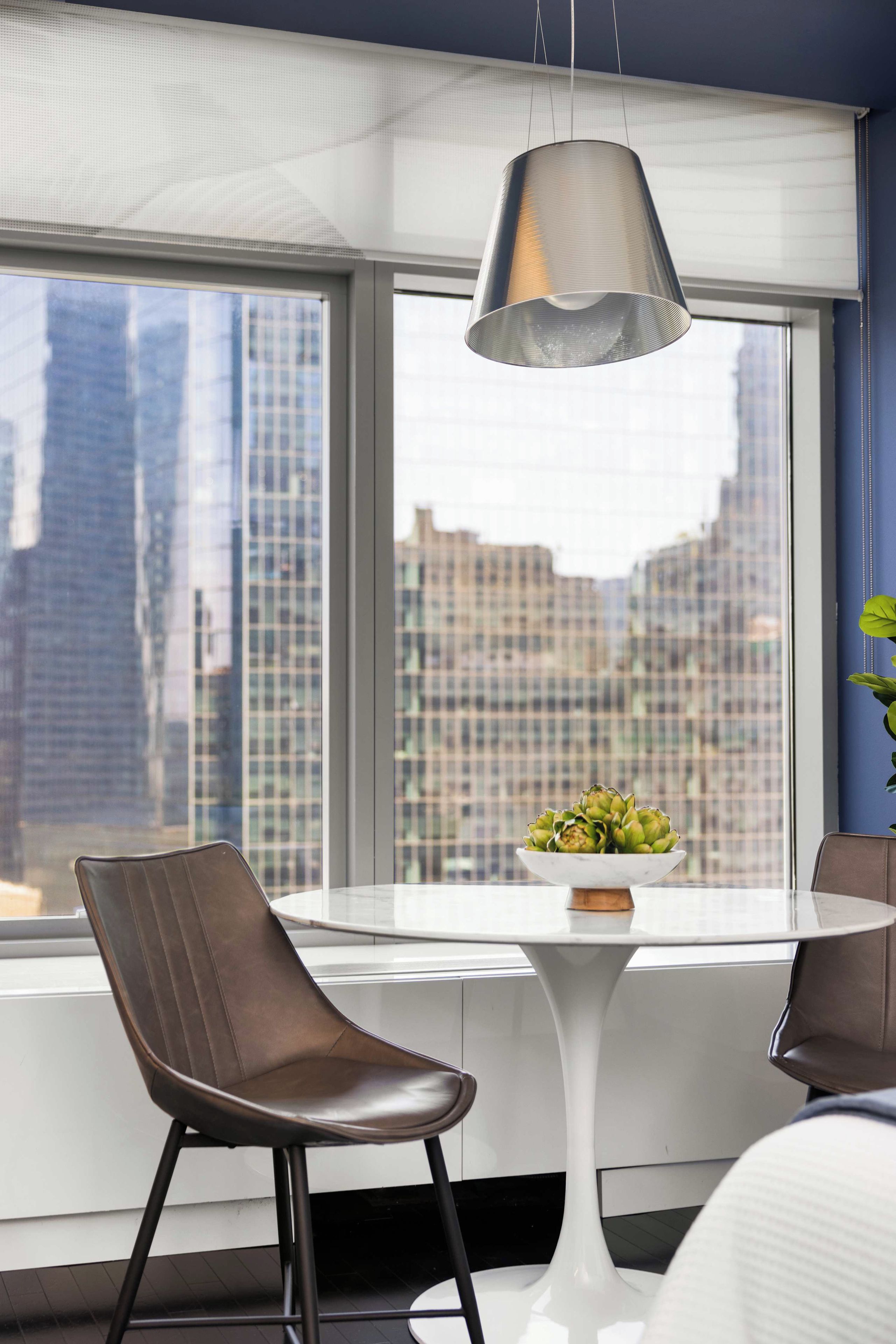 A modern dining area with a white table, two leather chairs, a hanging pendant light, and a view of city buildings through large windows.