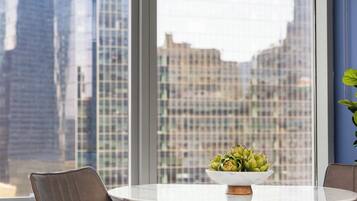A modern dining area with a white table, two leather chairs, a hanging pendant light, and a view of city buildings through large windows.