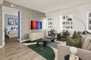 Ein modernes Wohnzimmer mit einer Couch, Sesseln, einem Couchtisch und einem Fernseher. Eine Tür führt in einen anderen Raum, und ein Fenster mit Jalousien ist vorhanden.