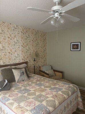 Ein Schlafzimmer mit einem Bett, einem Deckenventilator, einem Nachttisch, einer Lampe und einem gerahmten Bild an der Wand.
