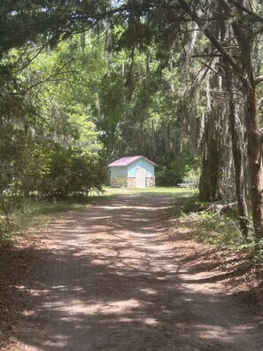 Ein schmaler Feldweg führt zu einer kleinen Holzhütte mit rotem Dach, umgeben von dichten Bäumen und viel Grün.