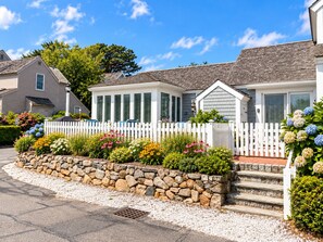 A residential house with a white picket fence, a stone retaining wall, and a variety of colorful flowers in the front yard. - A Storybook Cape Cod Cottage by the Sea (Mashpee)