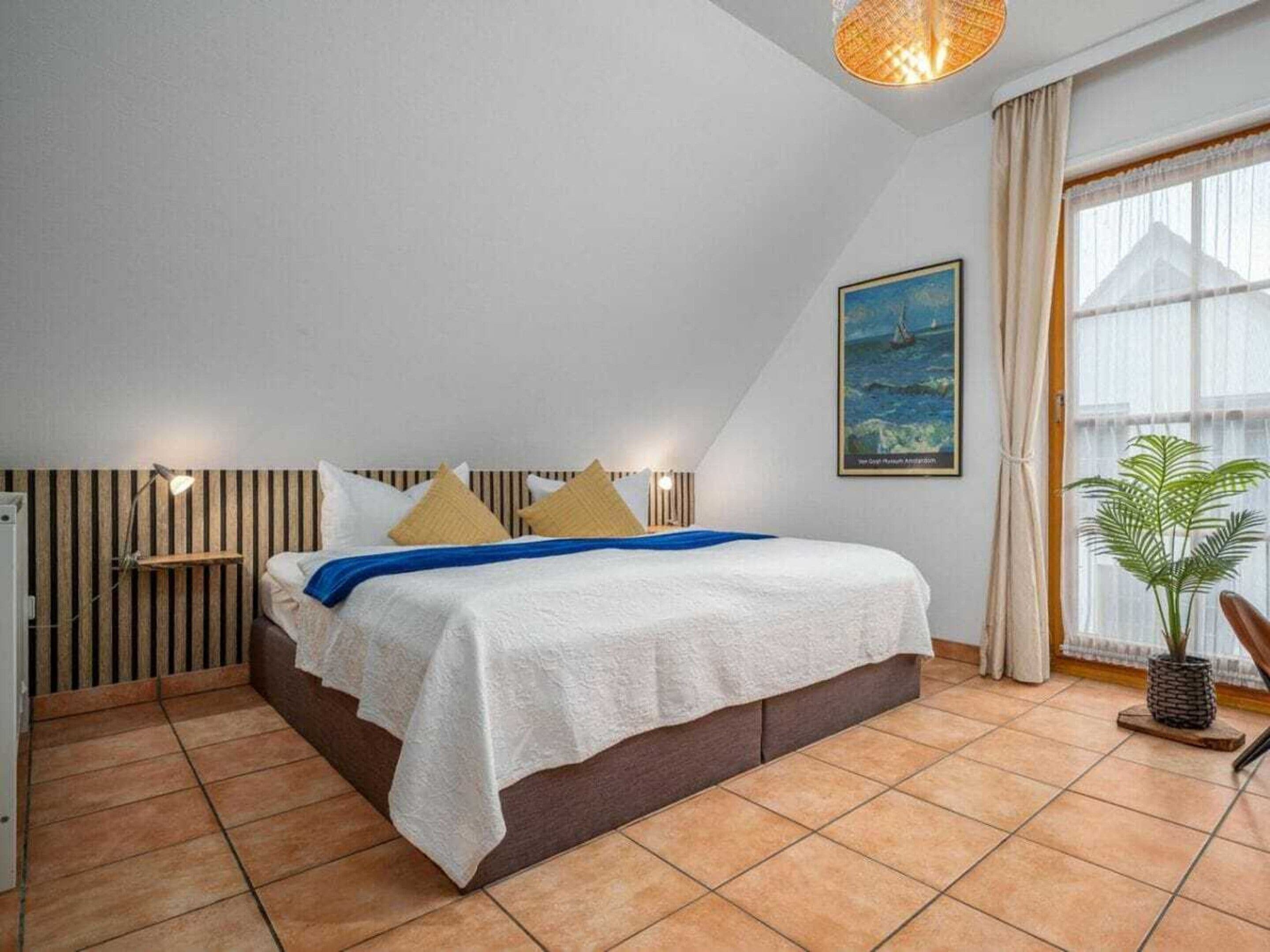 Ein Schlafzimmer mit einem Bett, einem Nachttisch, einer Lampe, einem Fenster mit Vorhängen, einer Pflanze und einem Bild an der Wand.