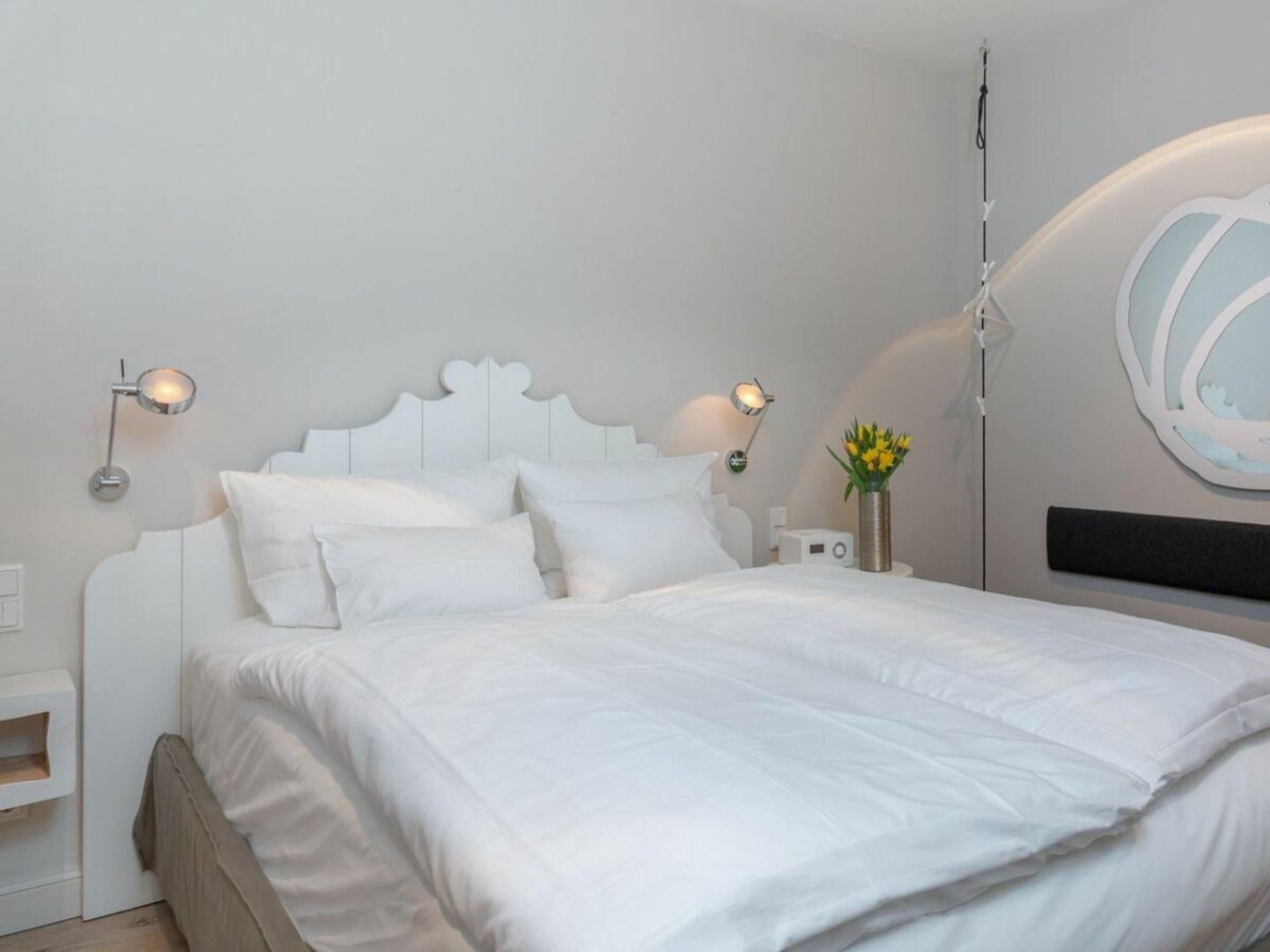 A bedroom with a white bed, two bedside lamps, a nightstand with a radio, and a wall-mounted mirror.