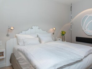 Un dormitorio con cama blanca, dos lámparas de noche, una mesita de noche con radio y un espejo montado en la pared.