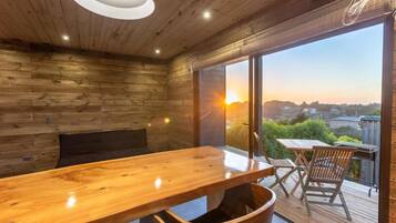 A wooden interior with a large table and chairs, a bench, and a view of the sunset through large windows.