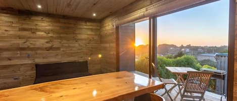 A wooden interior with a large table and chairs, a bench, and a view of the sunset through large windows.