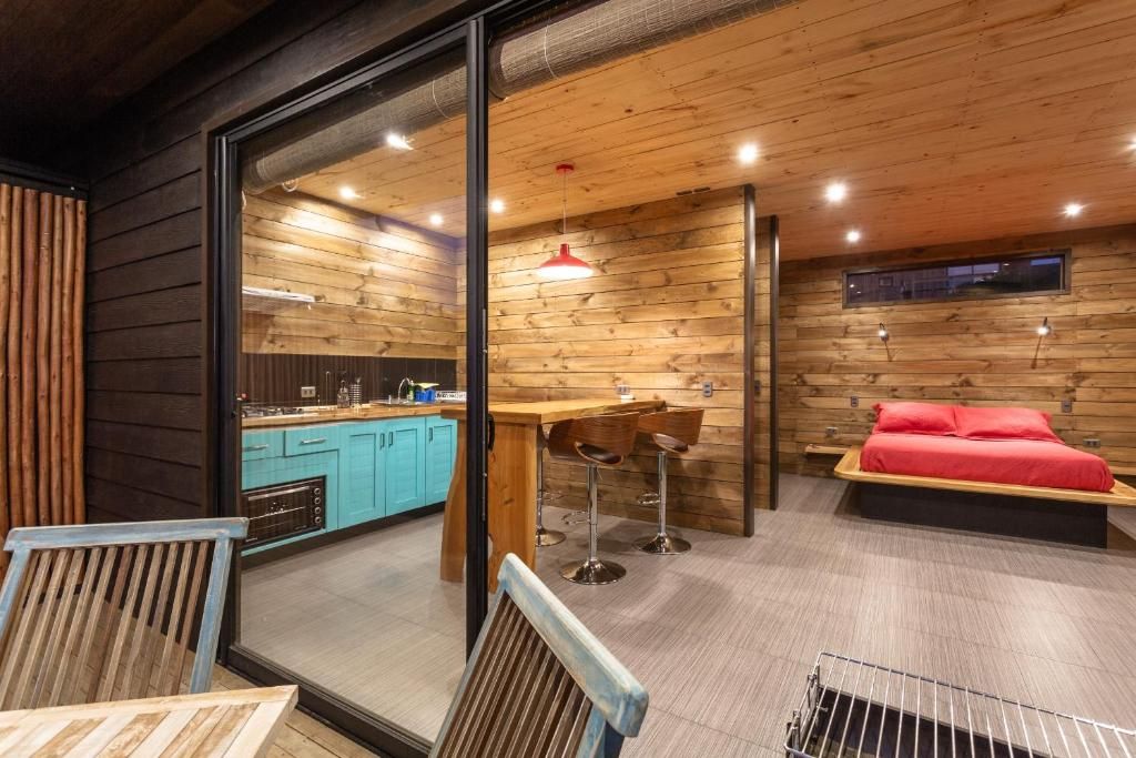 A modern interior with a wooden ceiling, a kitchen area, a dining space with stools, and a bed with red bedding.