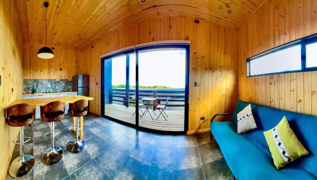 A wooden interior with a sofa, a dining area with stools, a kitchen, and a large sliding door leading to an outdoor deck.