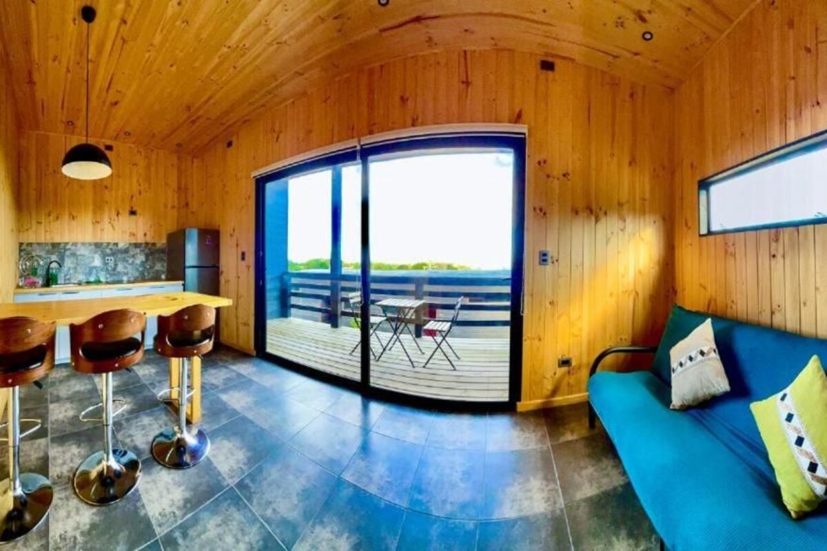 A wooden interior with a sofa, a dining area with stools, a kitchen, and a large sliding door leading to an outdoor deck.