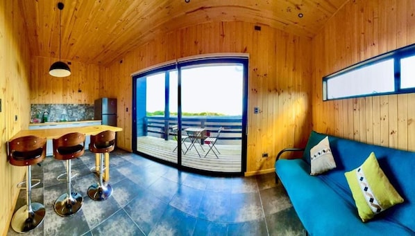 A wooden interior with a sofa, a dining area with stools, a kitchen, and a large sliding door leading to an outdoor deck.