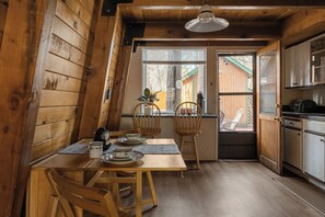 A wooden interior with a dining table set for two, a kitchen area, and a view through a window. - Enchanting 2BR Cabin with Private Spa • Big Bear Getaway (Big Bear)