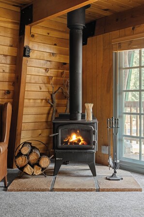 A wood-burning stove in a cabin with a wooden interior, a chair, and a window. - Enchanting 2BR Cabin with Private Spa • Big Bear Getaway (Big Bear)