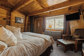A cozy cabin bedroom with a bed, dresser, bench, and a TV mounted on the wall. - Enchanting 2BR Cabin with Private Spa • Big Bear Getaway (Big Bear)