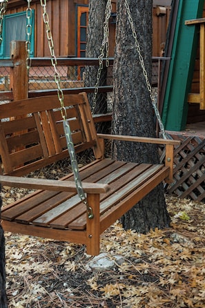 A wooden bench with metal chains, attached to a tree, surrounded by dry leaves and a wooden fence. - Enchanting 2BR Cabin with Private Spa • Big Bear Getaway (Big Bear)