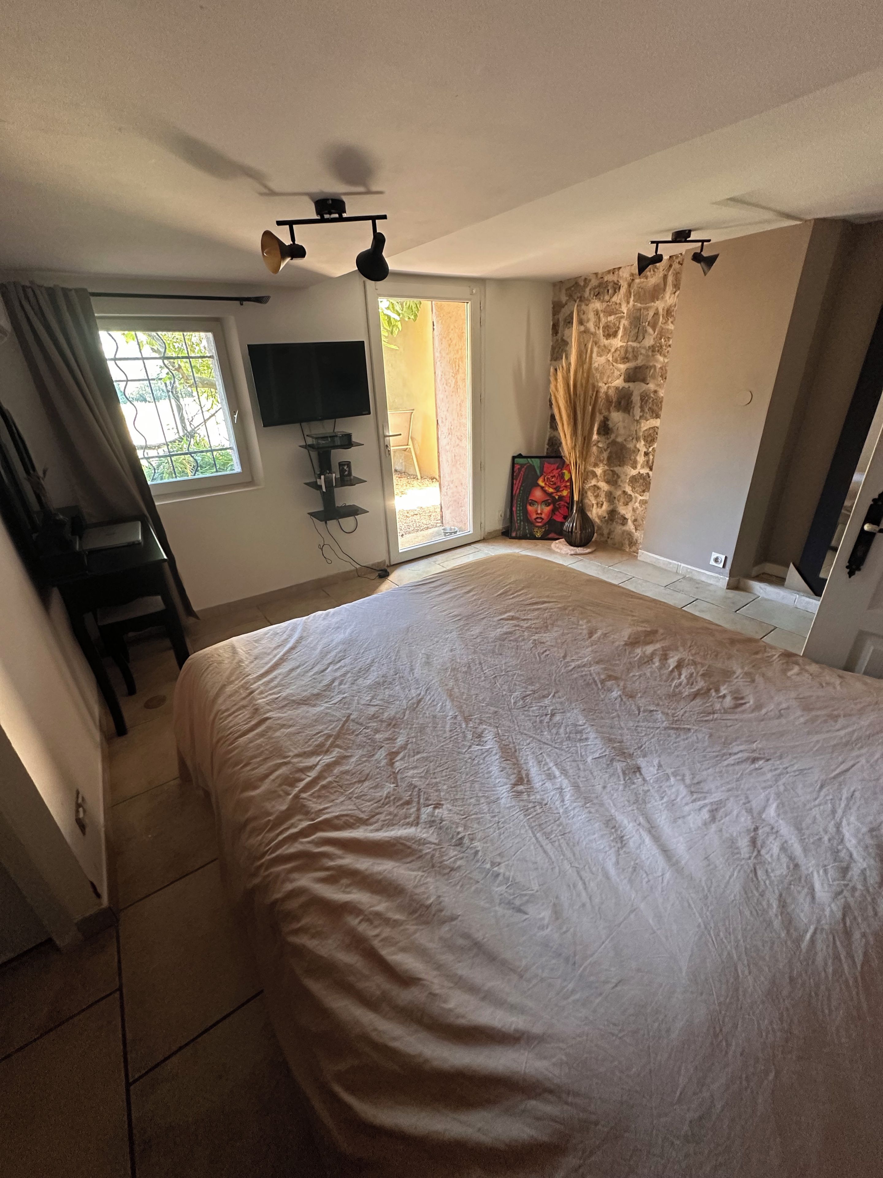 A bedroom with a large bed, a television, a window with curtains, a stone wall, and a door leading to an outdoor area.