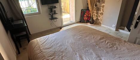 A bedroom with a large bed, a television, a window with curtains, a stone wall, and a door leading to an outdoor area.