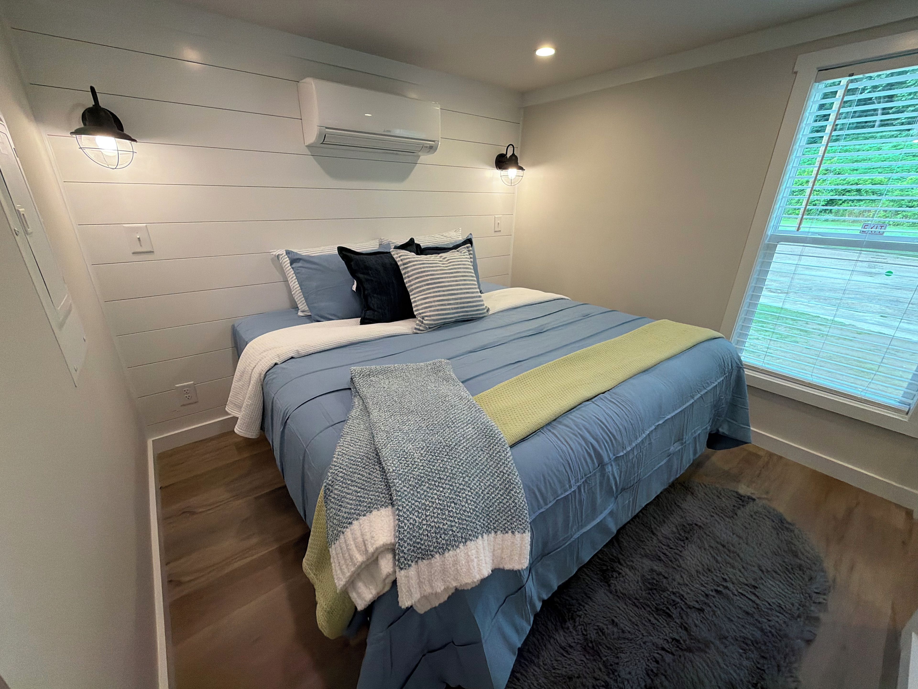 A bedroom with a bed, a window with blinds, an air conditioning unit, and a wall-mounted light fixture.