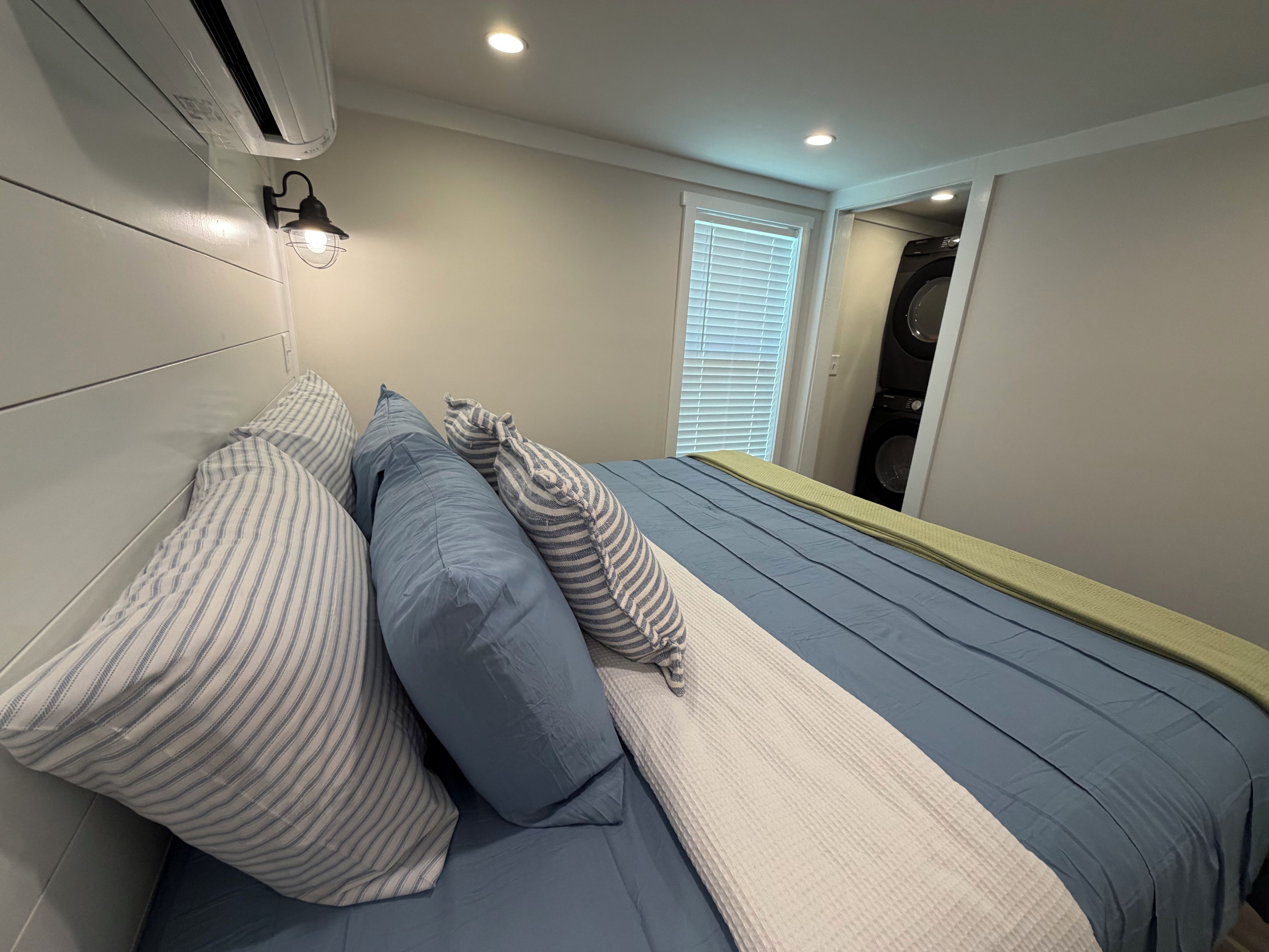 A bedroom with a bed, pillows, a wall-mounted air conditioner, a window with blinds, and a laundry machine in the background.