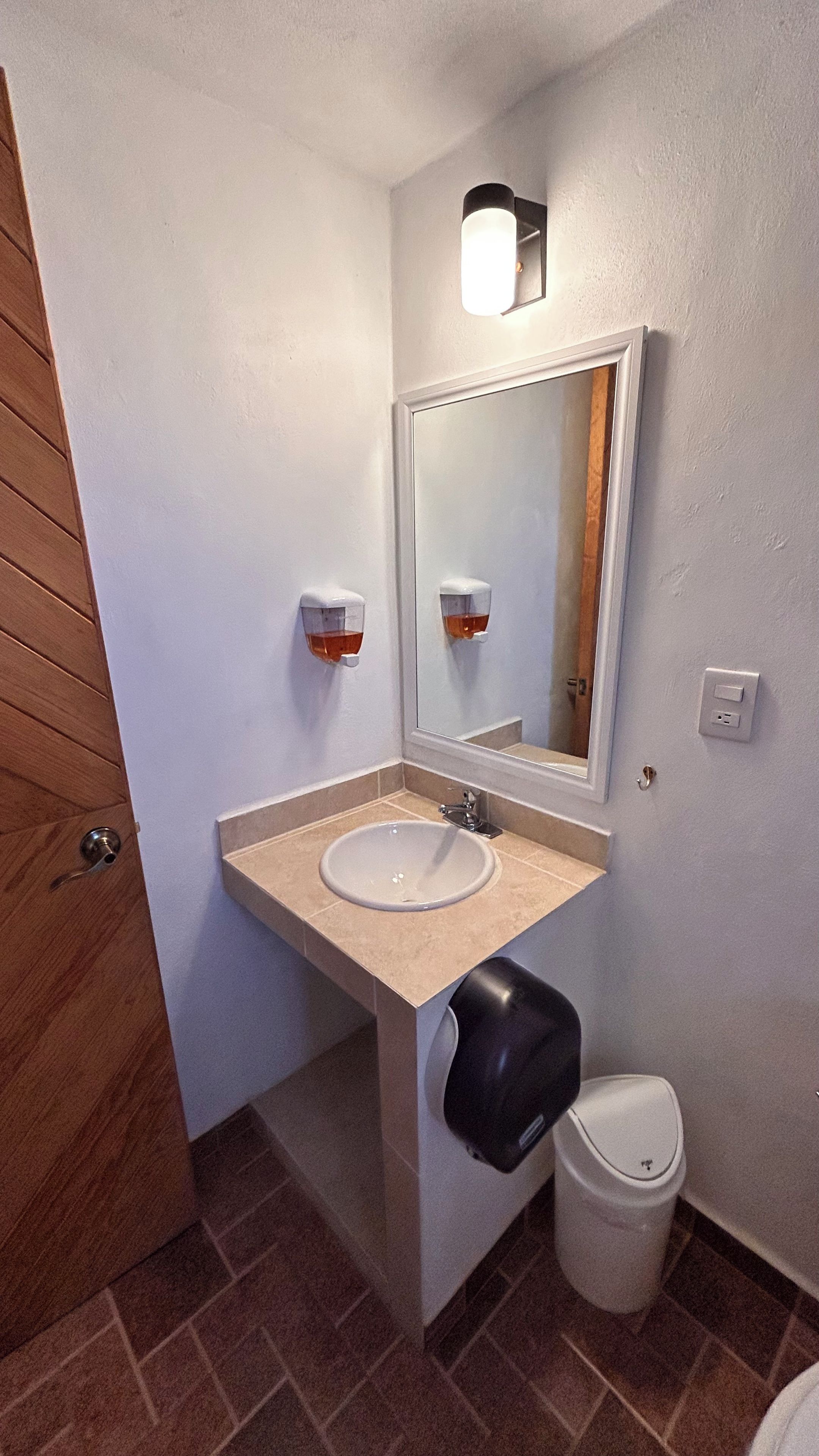 A bathroom with a sink, mirror, soap dispenser, and trash can.
