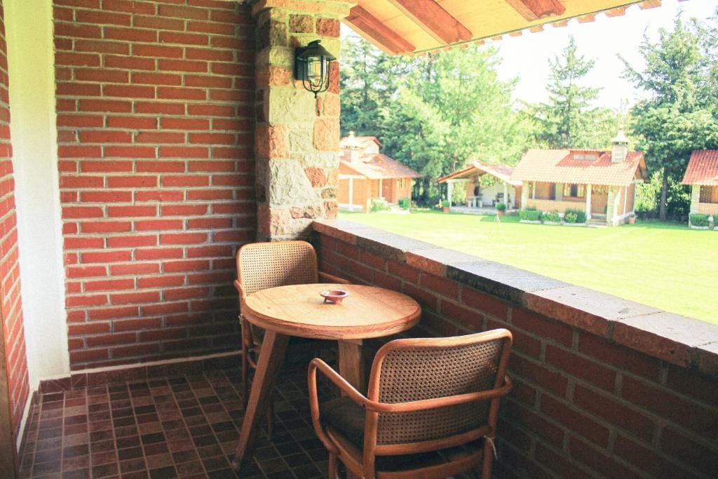 A covered patio with a brick wall, two wicker chairs, a small round table, and a view of a grassy area with houses.