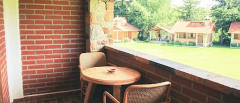 A covered patio with a brick wall, two wicker chairs, a small round table, and a view of a grassy area with houses.