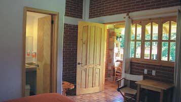 A room with a wooden door, a small table, a chair, and a window with a view of greenery.