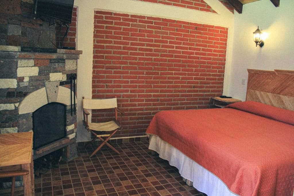 Una habitación con pared de ladrillo, chimenea, cama con manta roja, mesa de madera y silla plegable.
