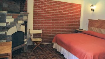 Una habitación con pared de ladrillo, chimenea, cama con manta roja, mesa de madera y silla plegable.