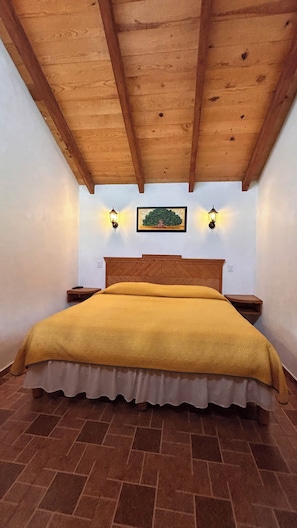 A bedroom with a wooden ceiling, a bed with a yellow bedspread, two bedside lamps, and a wall-mounted painting.