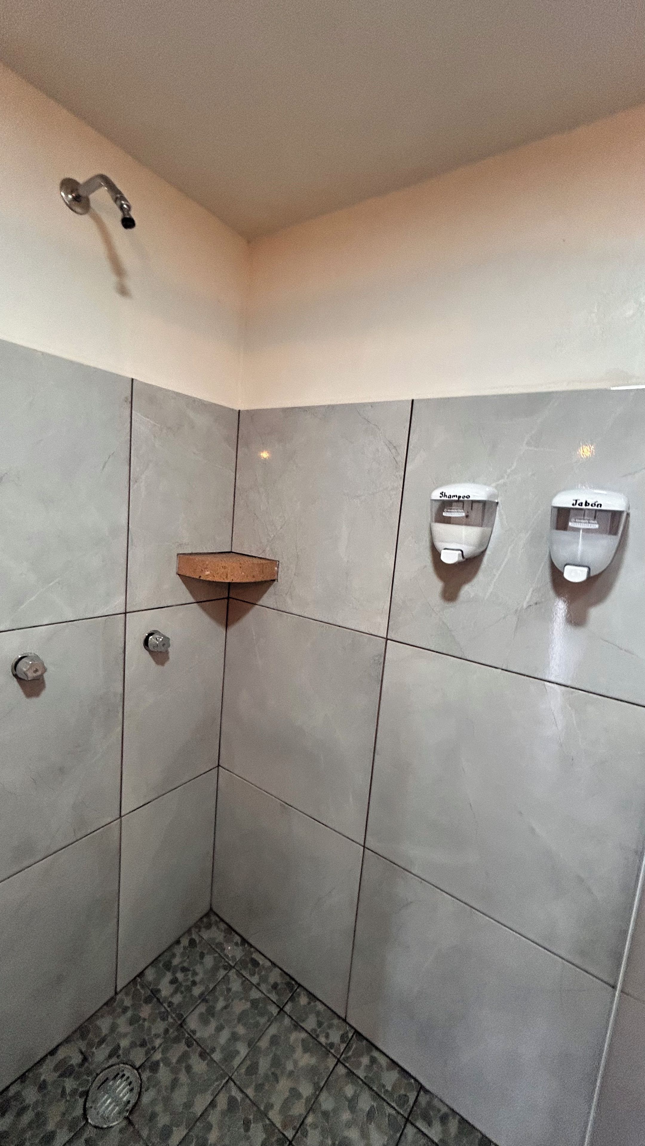 Un baño con azulejos grises, una ducha, un lavamanos con dispensadores de jabón y un estante de madera.