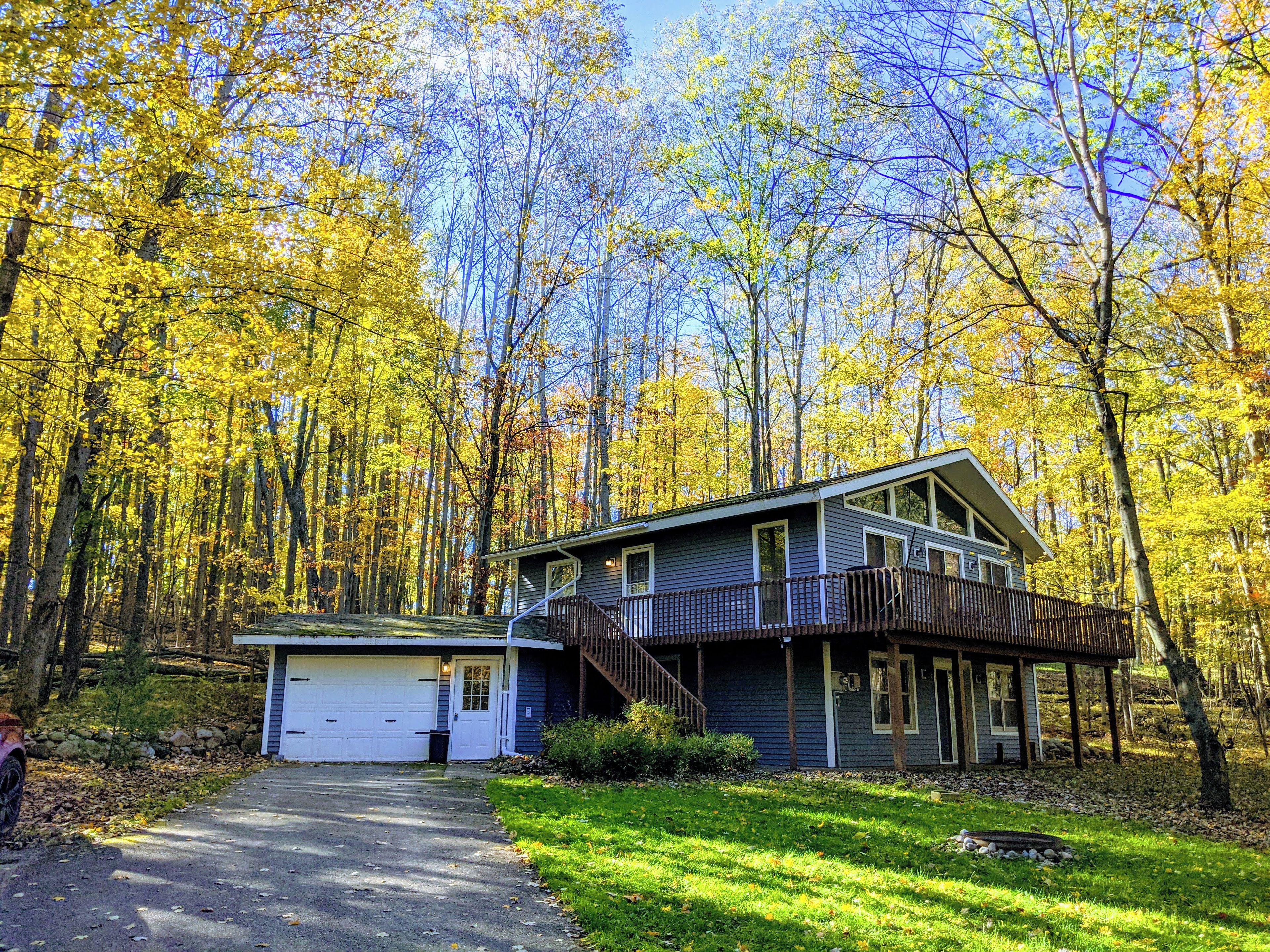 Une maison à deux étages avec un garage, entourée d’une forêt aux couleurs automnales.