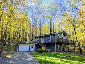 Une maison à deux étages avec un garage, entourée d’une forêt aux couleurs automnales.