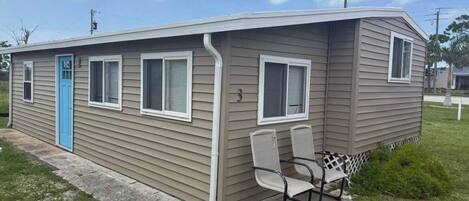 Una casa móvil de un piso con puerta azul, dos ventanas con contraventanas y un pequeño porche con dos sillas.