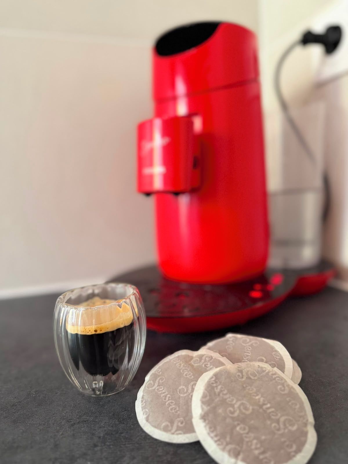 Una cafetera roja, una taza de vidrio con café y dos posavasos con texto.