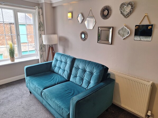 A teal sofa in a room with a radiator, a window with curtains, and various wall decorations. - Old Coach House Apartment (Scarborough)