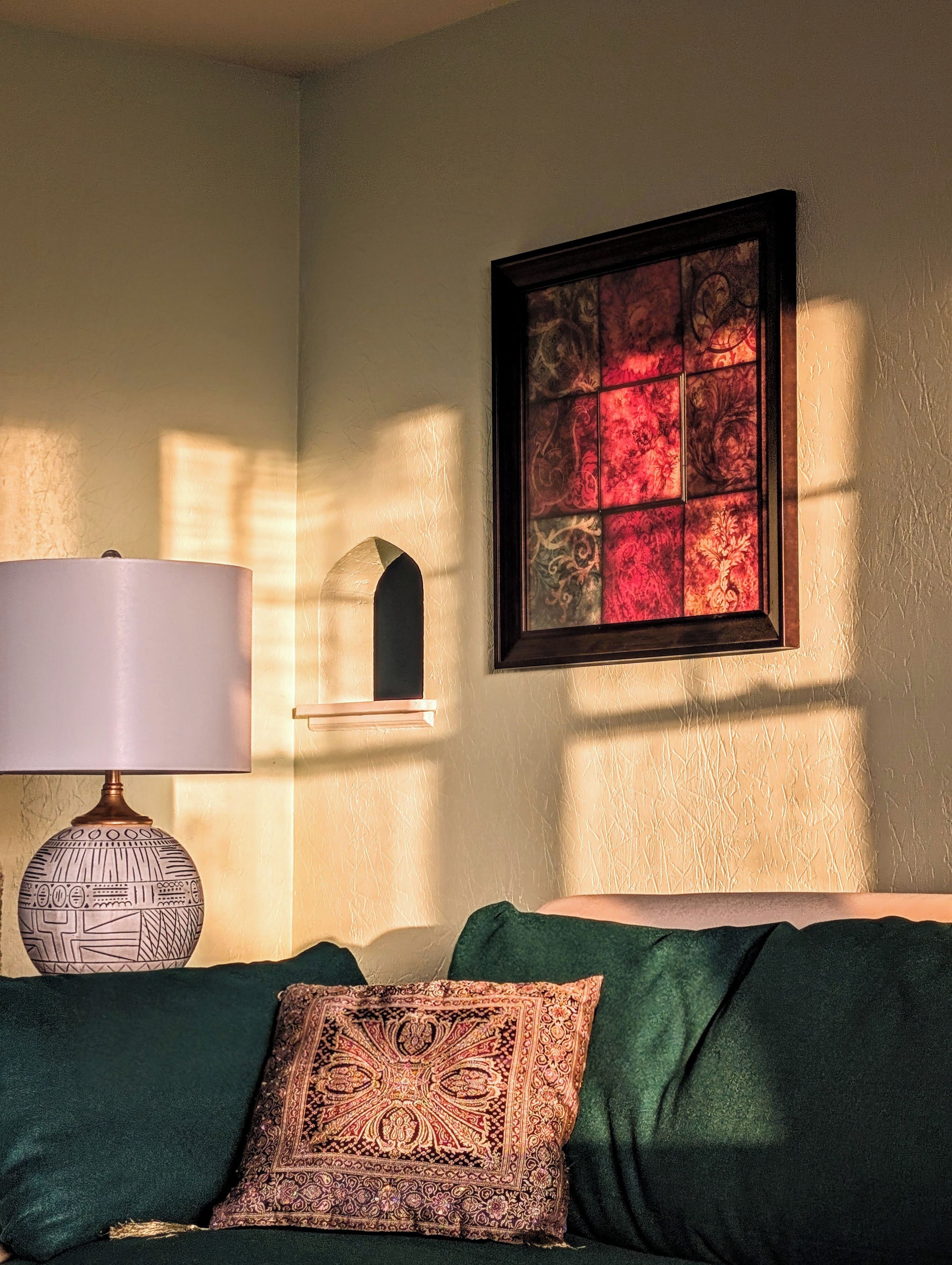 A room with a green sofa, a patterned throw pillow, a wall-mounted shelf with a decorative item, and a framed artwork with a red and gold pattern.