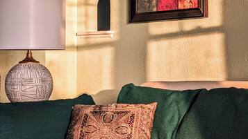 A room with a green sofa, a patterned throw pillow, a wall-mounted shelf with a decorative item, and a framed artwork with a red and gold pattern.