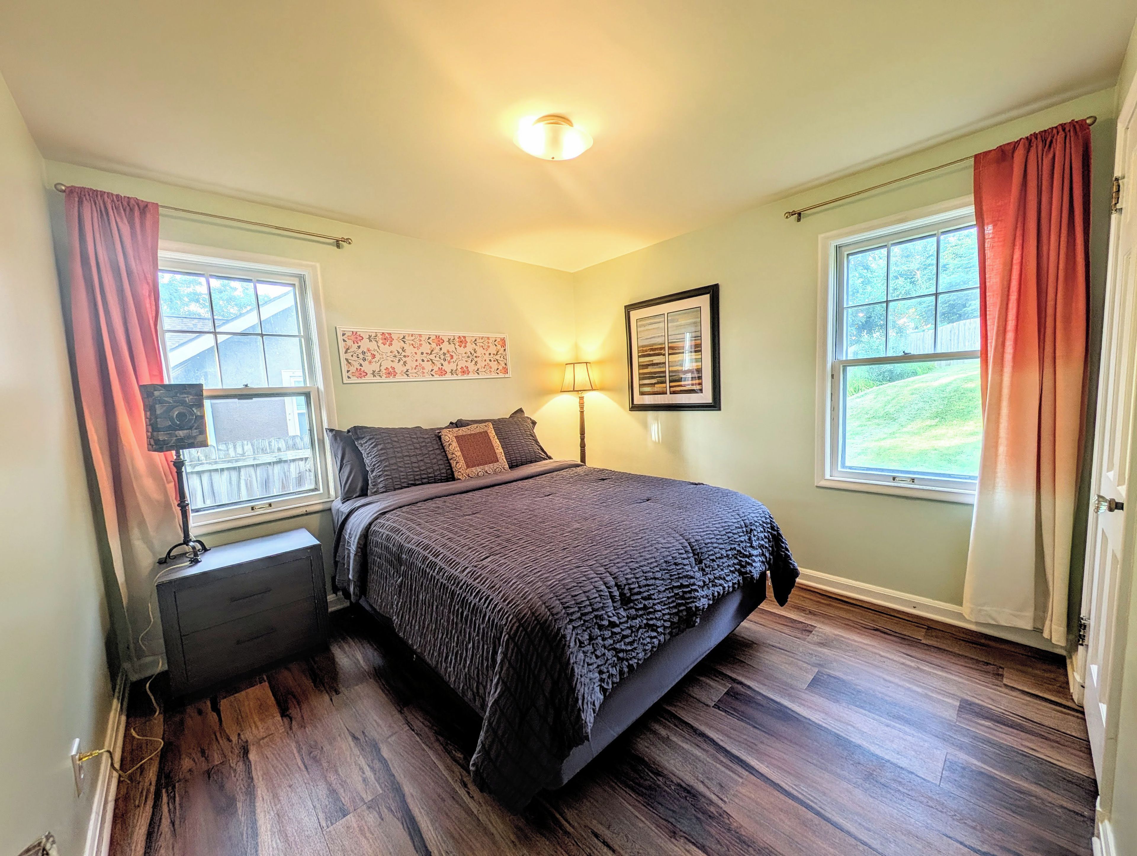 A bedroom with a bed, nightstand, lamp, and two windows with curtains.