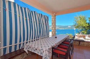 Um terraço com mesa e cadeiras, uma cortina listrada e vista para o mar e montanhas.