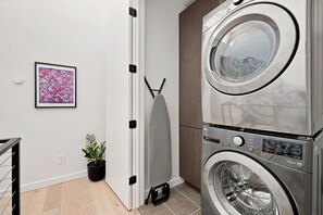 Een wasruimte met een gestapelde wasmachine en droger, een potplant en een lijst met abstracte kunst aan de muur.